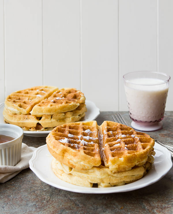 waffles stacked on two plates with a glass of milk and syrup