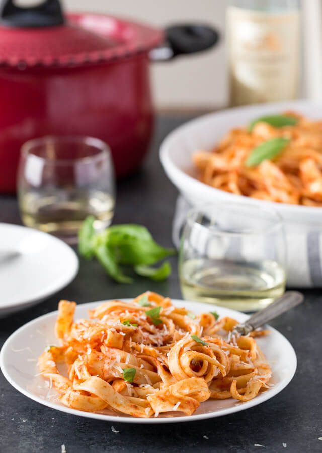 pasta on a white plate with wine glasses and big pasta dish in the background